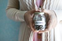 A pair of hands holding a small mason jar filled with coins and a label on the jar that reads: "Savings"