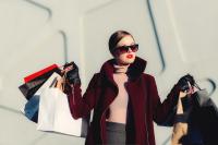 A white woman is dressed very chic in a red coat and dark sunglasses. She is holding numerous paper shopping bags in both hands like she just finished a big shopping spree.