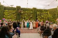 Dana and Anya raise their arms triumphantly during the wedding ceremony. They are surrounded by loved ones waving pride flags and a green, leafy background.