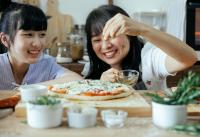 Two Asian women sprinkling seasoning on an uncooked pizza. Sustainable kitchen products.