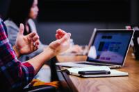 person sitting at a table in front of a laptop, gesturing with their hands, a woman sits in the background, root & leaf specializes in marking consulting