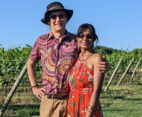 Swati & Mark standing together against a green landscape. Swati is wearing a bright orange dress and they are both smiling.