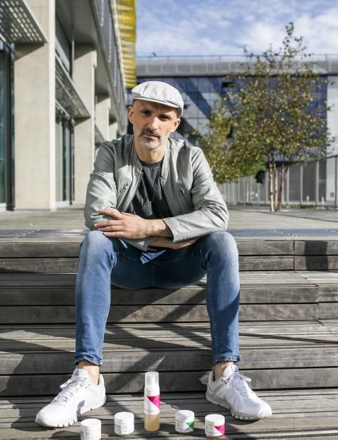 Bo, a white man wearing a newsboy cap, sits on wooden stairs outside with a line of skincare products in front of him.