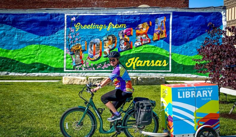 A woman is riding a green bike with a box attached to the back that reads "Topeka & Shawnee County Public Library." She is in front of a mural that says "Greetings from Topeka, Kansas."