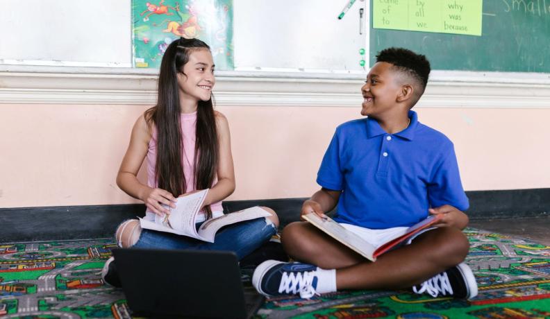 An elementary school aged girl and boy sit on the floor with books in their laps. They are smiling at each other.