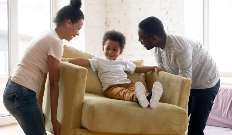 A Black mom and dad pick up an armchair with a 4 year old kid sitting in it. They're all smiling and laughing together.