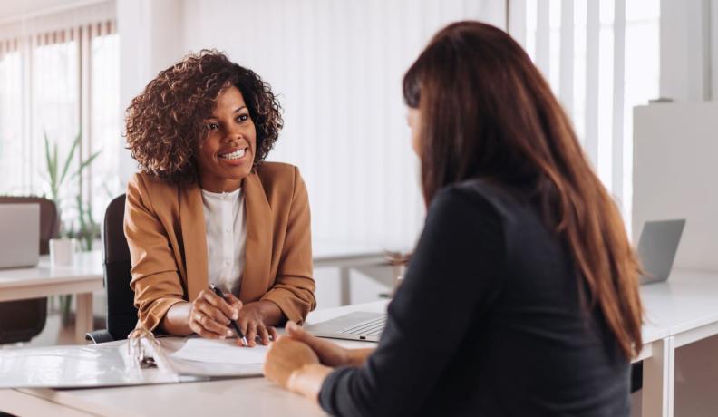 Black woman financer wearing a brown blazer and shoulder-length hair consulting another woman. 