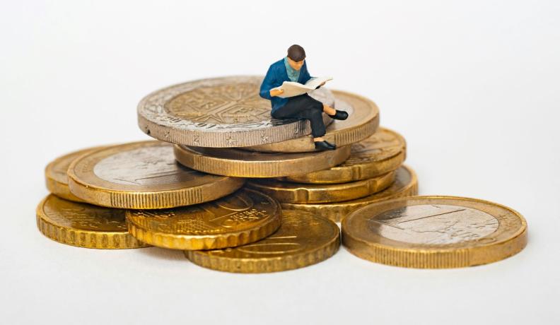 miniature figure of a person in a blue jacket sitting on top of a stack of gold coins.