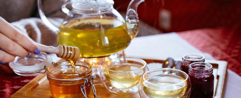 A wooden tray with tea in a glass kettle, jars of jam, and an open mason jar of honey. A hand sticks out from the left holding a honey dipper. Sustainable honey products