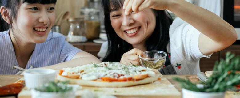 Two Asian women sprinkling seasoning on an uncooked pizza. Sustainable kitchen products.