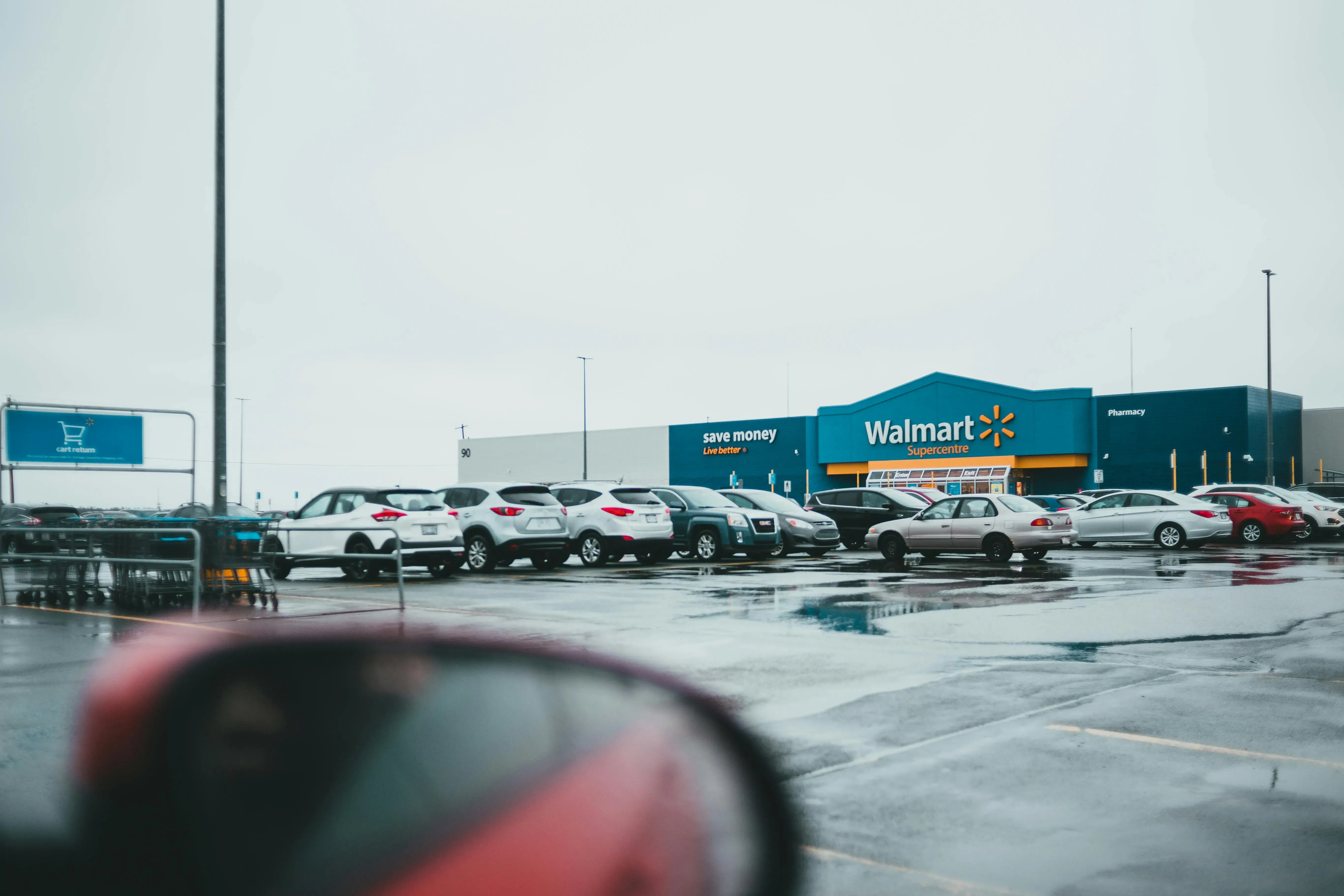 A picture of a Walmart store in the distance, taken from a car in a large parking lot. The car side mirror is visible in the forground.