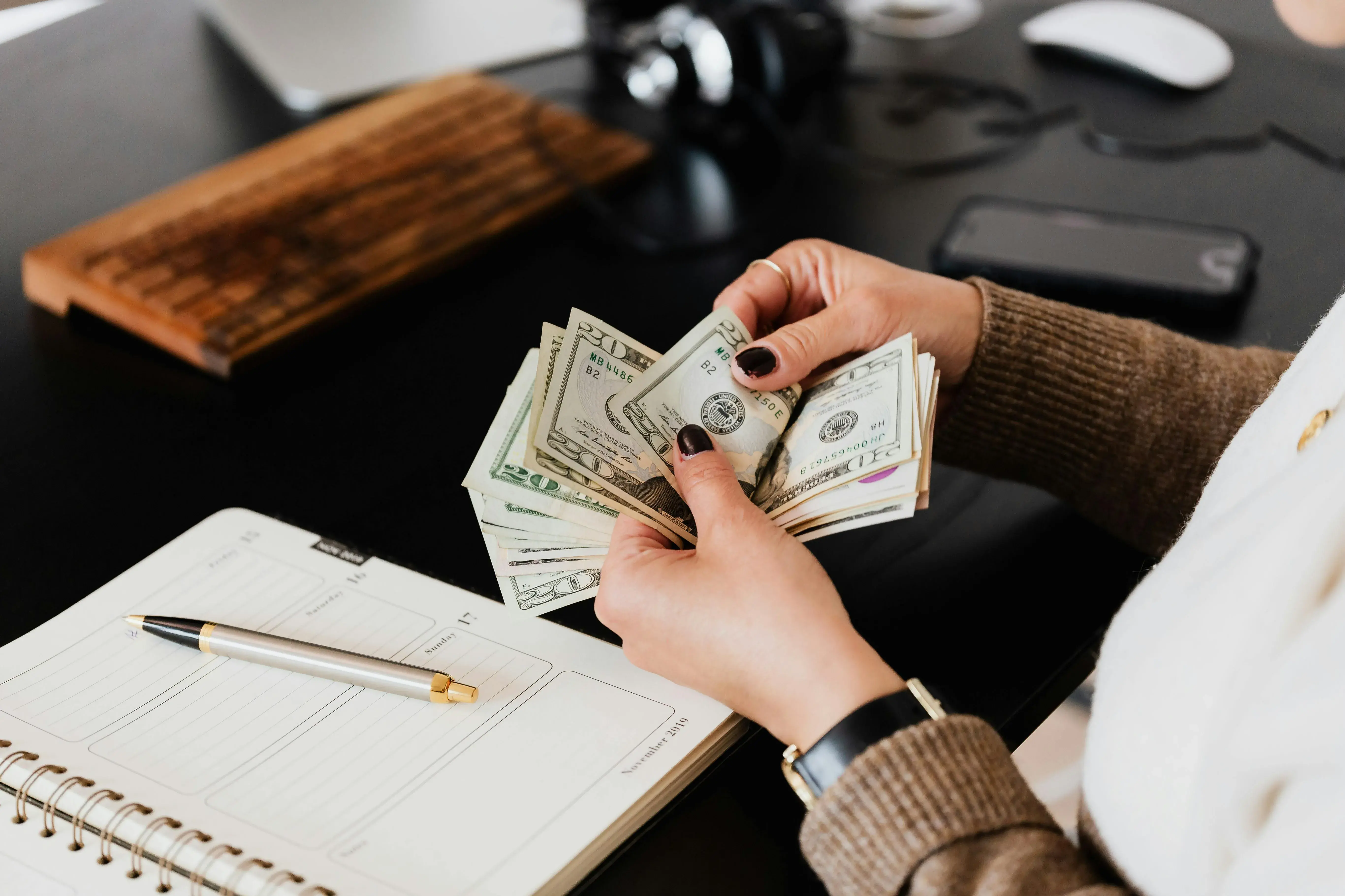 a woman's hands counting cash with a notebook on a desk in front of her