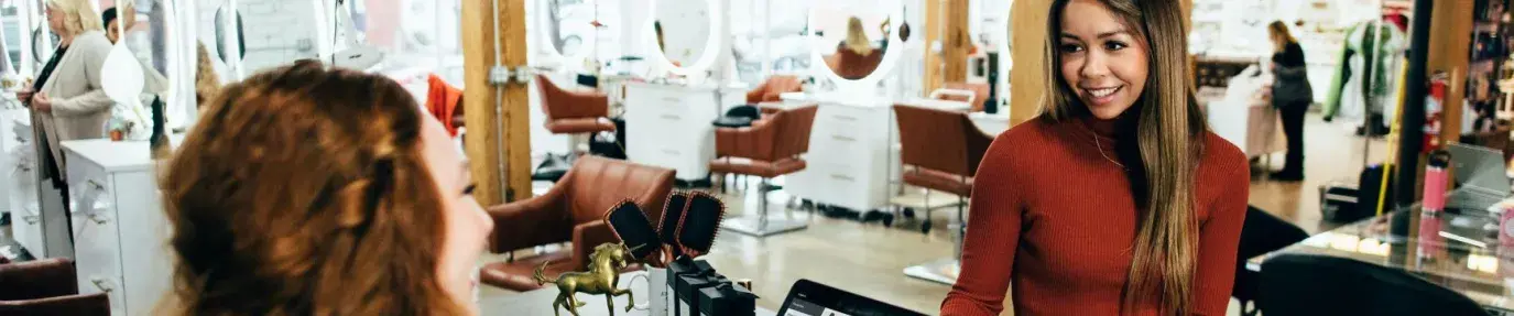 red-haired salon worker with a big smile standing behind counter accepting payment from a woman customer in a red sweater with a big smile, businesses that do a sustainability audit are on their way