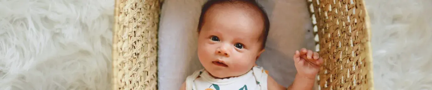 baby laying in crib wearing a onesie with oranges on it, looking curiously at the camera. The crib is wicker and is on top of a white faux fur rug. The mattress still matters for everyone.