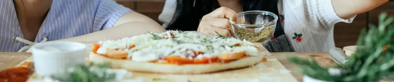 Two Asian women sprinkling seasoning on an uncooked pizza. Sustainable kitchen products.