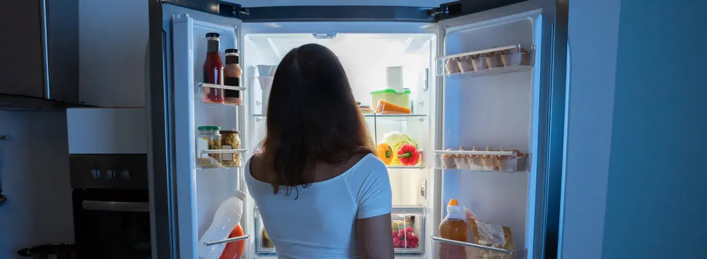 Image: woman looking into open fridge at night. Title: Climate-Friendly Fridges That Are Truly Cool