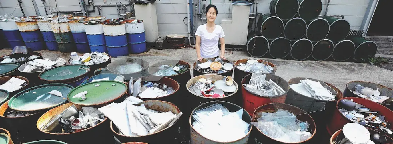 Image: woman standing with full recycling containers. Topic: Americans are bad at recycling. Here's How the World Does It Better.