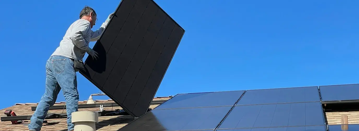 A man stands on a rooftop installing solar panels, he carries one with several others already laid out on the roof.