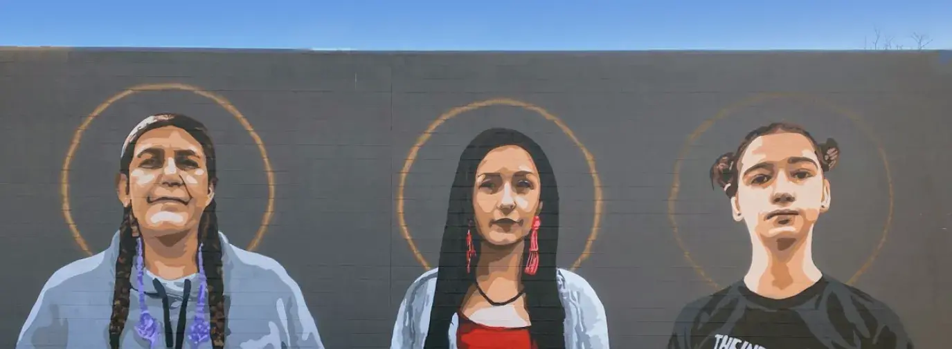 Mural painted on the outside of Native American Bank. There are three women of different ages standing side by side with halos around their heads to represent what Native American Bank honors and celebrates.