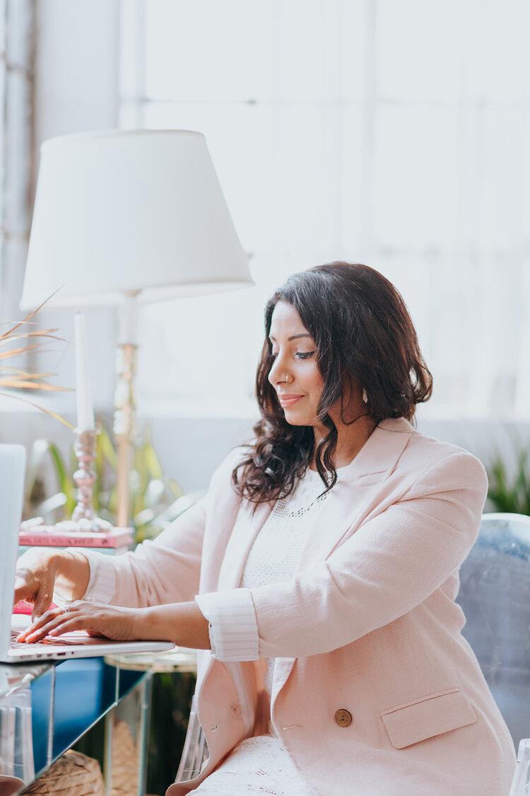 Adila Cokar, an Indian woman wearing a pale pink blazer sits at a table typing on a Mac laptop. The Good Tee.