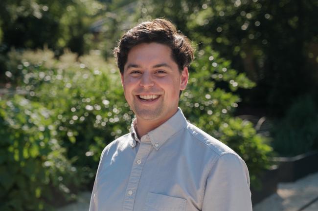 Alex Lassiter, a white young man with brown hair and a wide smile on his face. He stands outside in front of some large, green bushes.