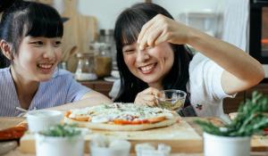 Two Asian women sprinkling seasoning on an uncooked pizza. Sustainable kitchen products.