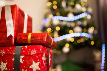 a stack of three presents wrapped in red, shiny paper are sitting in front a Christmas covered in yellow and blue lights.