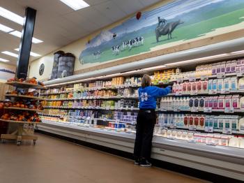 A woman in a blue trader joe's employee shirt organizing the shelves in the dairy section.