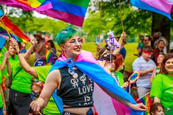 Happy person with rainbow makeup wearing a trans flag as a cape in a parade of rainbow flags.