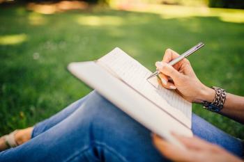 A woman's hand writes in a notebook resting on her knees. She's sitting in a grass field. Sustainable creative accessories.