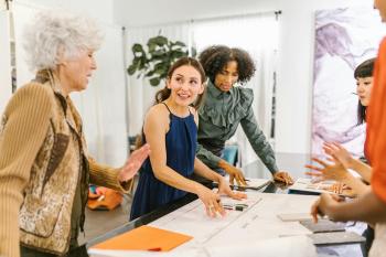 A group of women stand around a table covered in paper. They are actively discussing something. Women Business Owners.